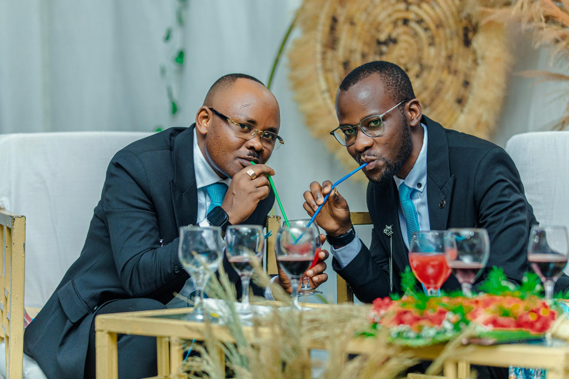 Zwei elegant gekleidete Herren in Anzügen genießen bei einer gesellschaftlichen Veranstaltung ein Getränk mit Strohhalm.